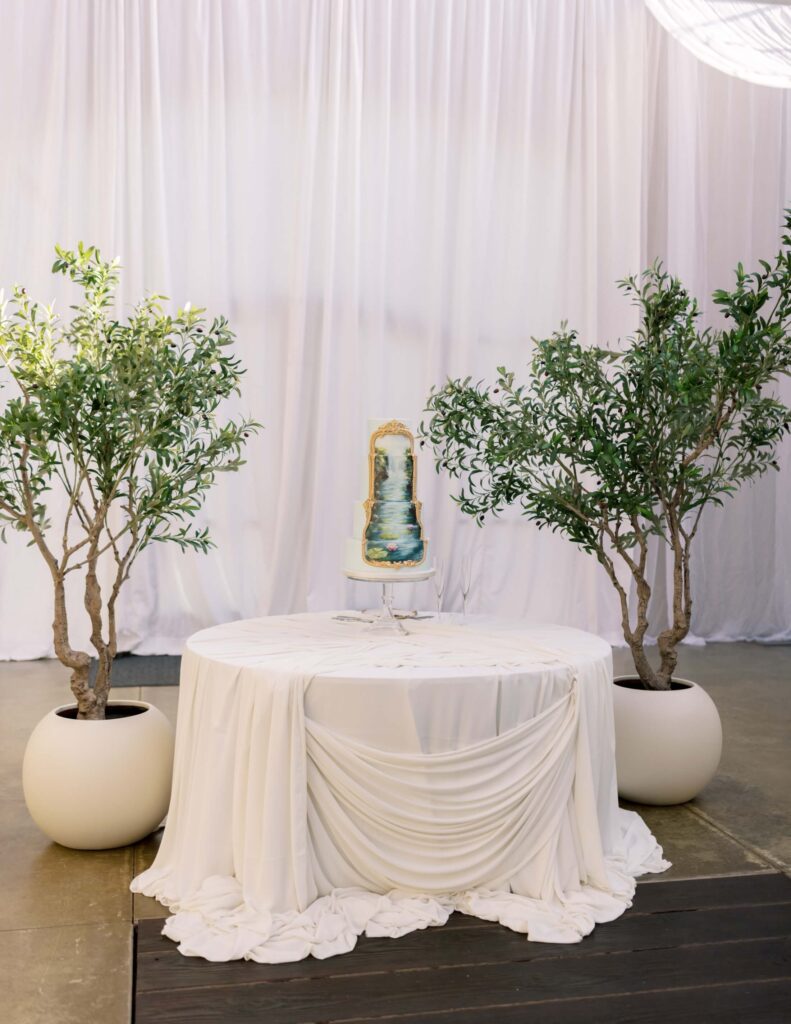 A wide view of a wedding cake display table featuring a four-tier cake with an original Impressionist painting reproduction by Alana Eden, beautifully flanked by two large potted trees against flowing white draped linens.
