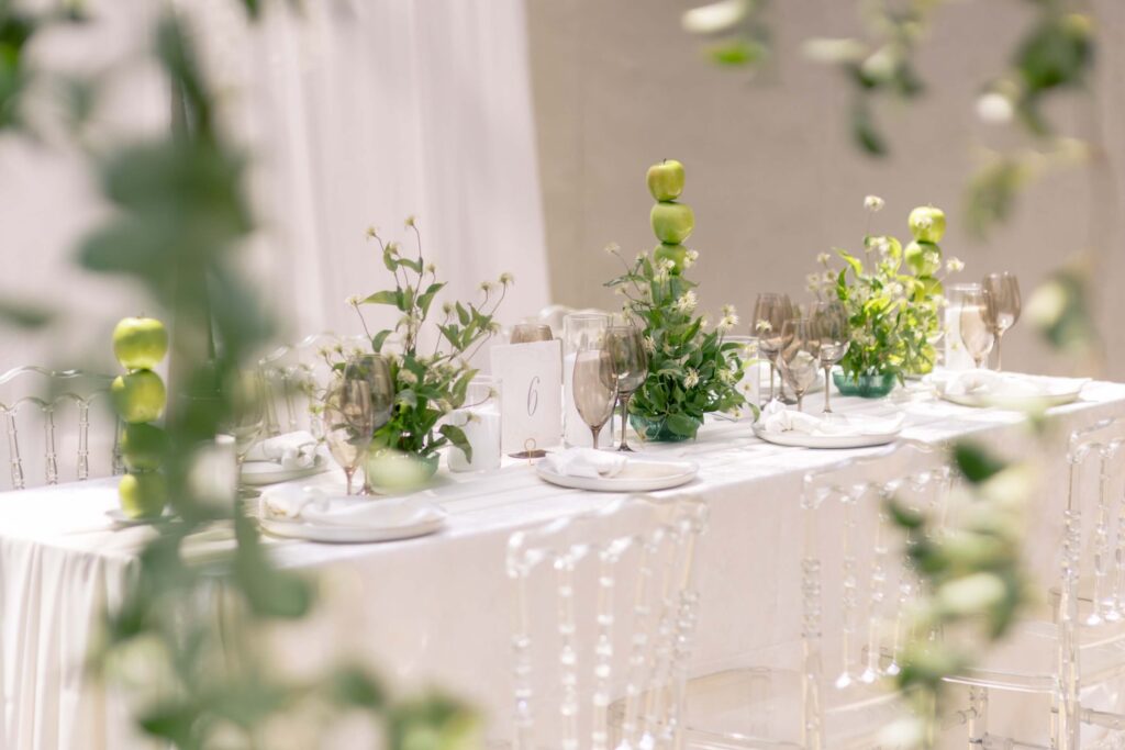 Fine art wedding reception table featuring acrylic ghost chairs, unique green apple centerpieces, and a custom Art Nouveau table number six card by Eden Hill Atelier, viewed through blurred foreground greenery.⁠