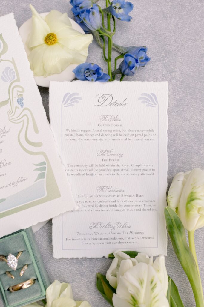 Vertical flat lay focusing on a deckled-edge wedding details card by Eden Hill Atelier featuring slate blue Art Nouveau design elements, styled with fresh blue delphiniums, white tulips, and a sage green velvet ring box.⁠