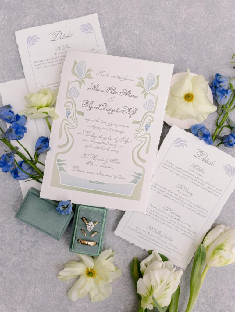 Flat lay of a custom Art Nouveau wedding invitation suite by Eden Hill Atelier featuring sage green and dusty blue botanical borders, deckled-edge paper, a green velvet ring box, and white flowers.
