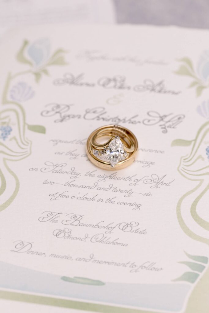 Close-up macro shot of gold wedding bands and a diamond engagement ring resting on a custom Art Nouveau wedding invitation by Eden Hill Atelier, featuring delicate sage green and dusty blue botanical line work.