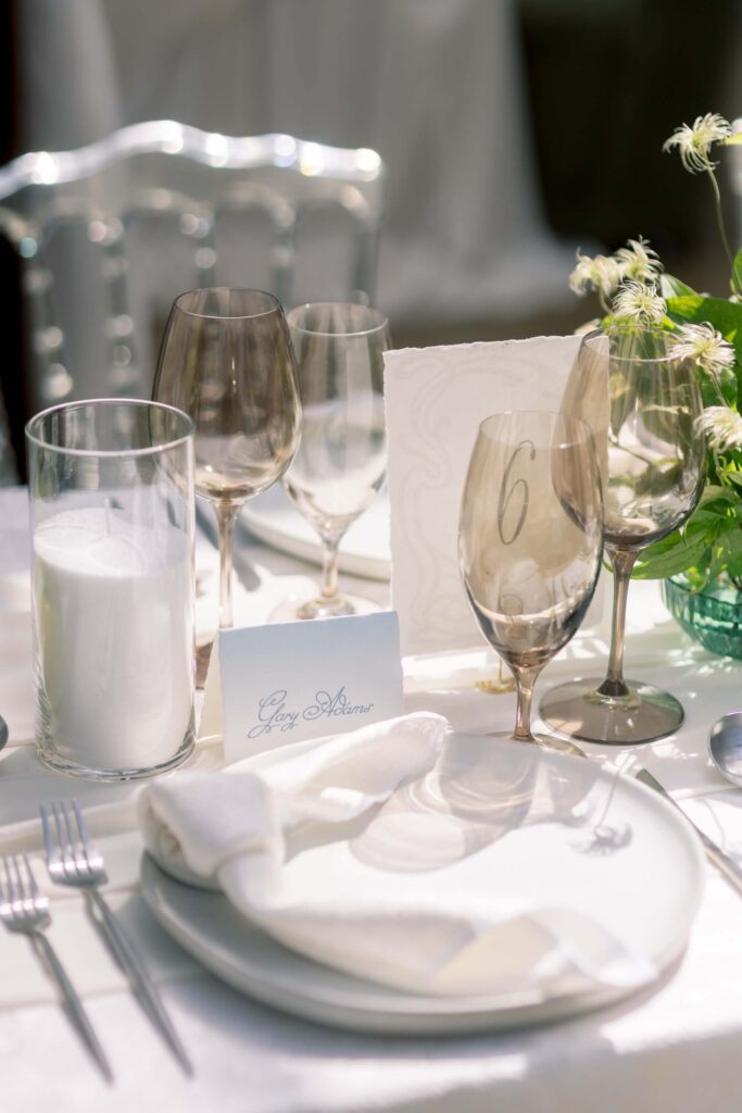 Close-up of a custom wedding place card with fluid script calligraphy reading Gary Adams, styled on a reception table setting with smoky tinted wine glasses, a white plate, and a clear acrylic chair.⁠