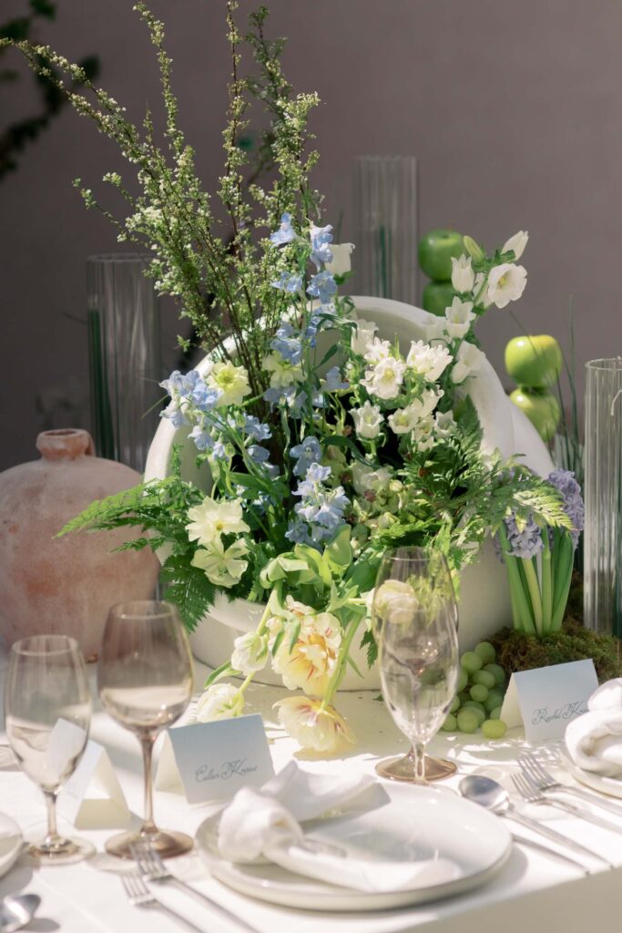 Fine art wedding reception tablescape highlighting custom script place cards by Eden Hill Atelier styled alongside a large white ceramic floral centerpiece, fresh green grapes, and tinted stemware.