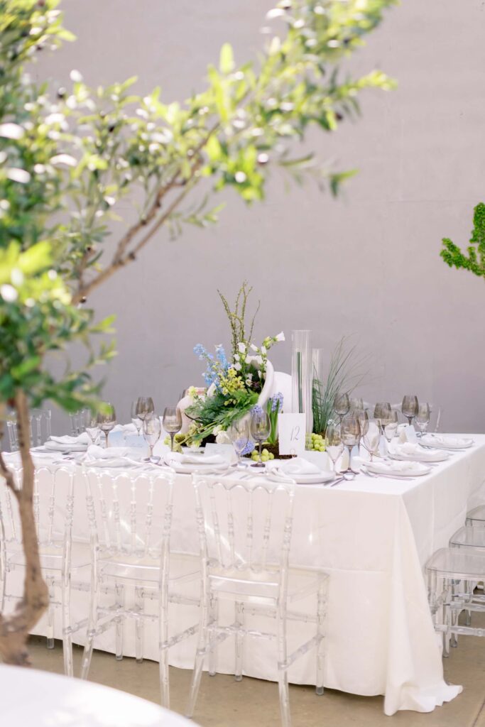 A fine-art wedding reception table setup featuring clear acrylic ghost chairs, crisp white tablecloths, tinted glassware, and a custom table number card by Eden Hill Atelier.