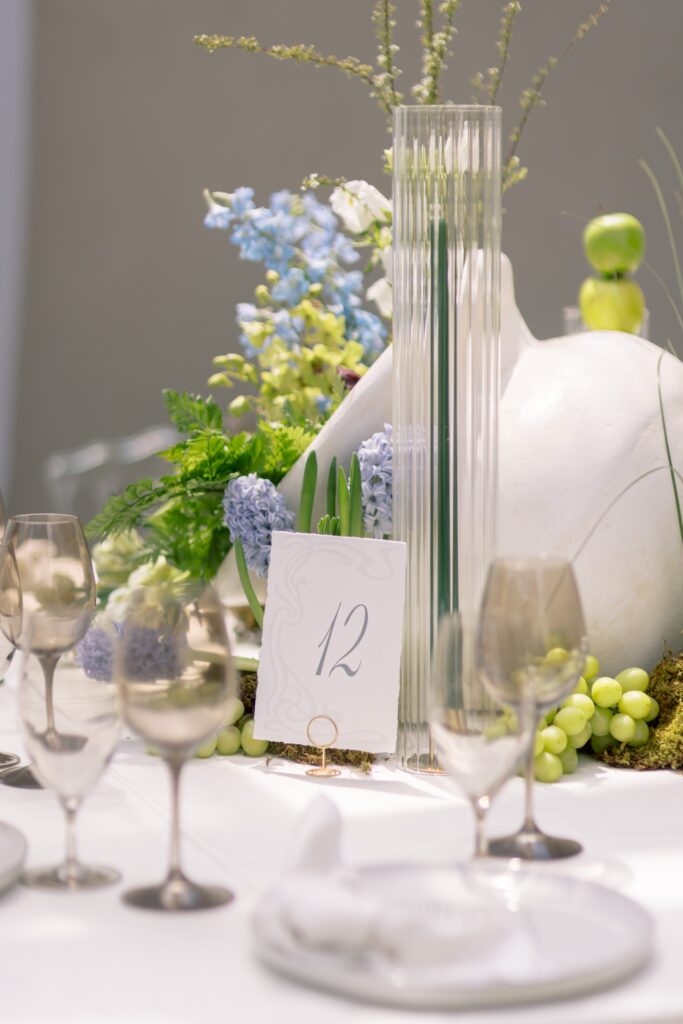 Custom wedding table number card displaying number 12 with a delicate Art Nouveau border, styled on a reception table centerpiece with a blue floral arrangement, a green taper candle, and smoked glassware.