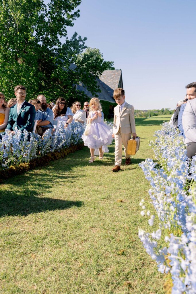 A ring bearer in a tan suit and a flower girl in a light purple dress walk down a grass wedding aisle lined with heavy blue delphinium floral installations at The Baumberhof.