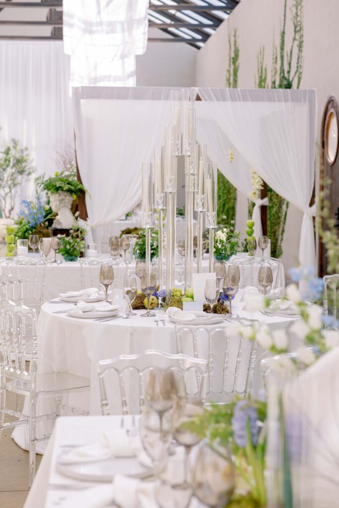 An indoor wedding reception setup at The Baumberhof showcasing a towering glass candelabra centerpiece with white taper candles on a round table styled with white linens and clear acrylic ghost chairs.