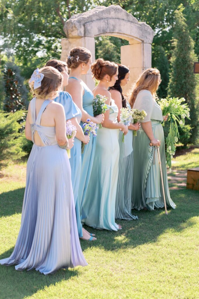 A side profile view of bridesmaids standing on a sunlit lawn in mismatched pastel blue, periwinkle, and sage green dresses during an outdoor ceremony at the stone arch at The Baumberhof.⁠