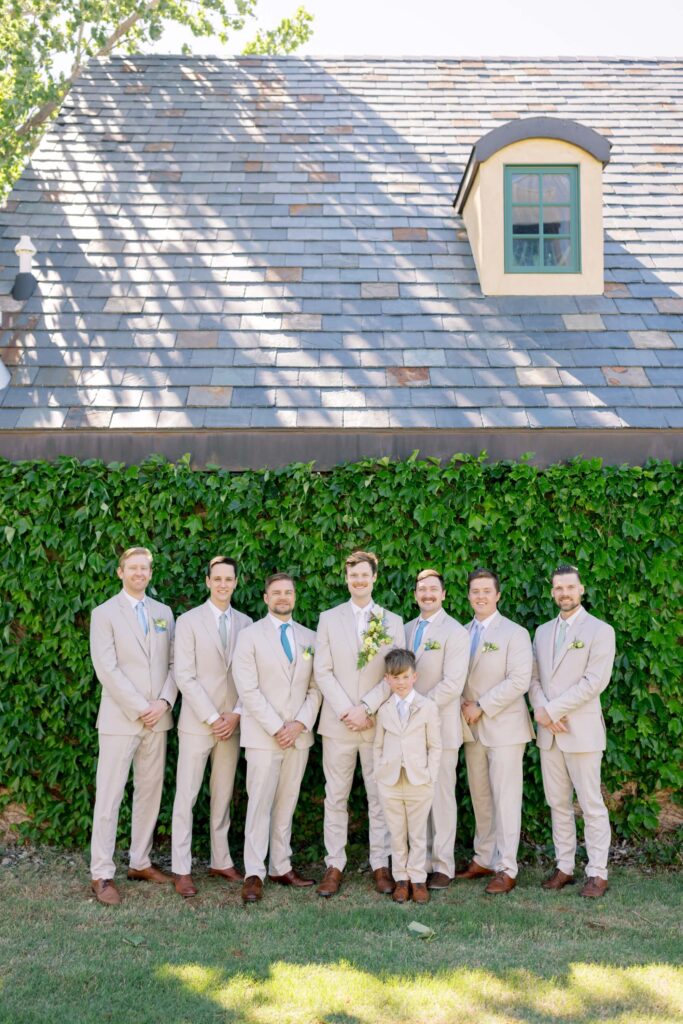 The groom, groomsmen, and a young ring bearer standing in a row in matching tan suits with periwinkle ties in front of a lush green ivy wall under a slate-shingled roof at The Baumberhof.⁠