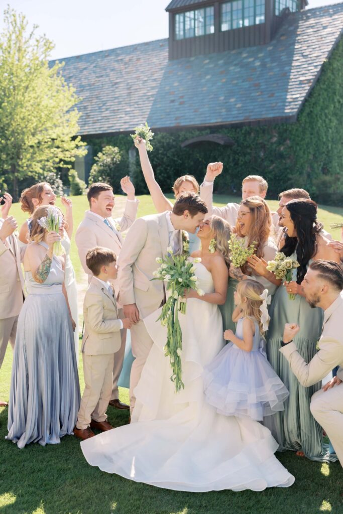 Bride Alana and groom Ryan kiss in the center of their cheering wedding party on the lawn at The Baumberhof, surrounded by bridesmaids in mismatched pastel gowns and groomsmen in tan suits.⁠