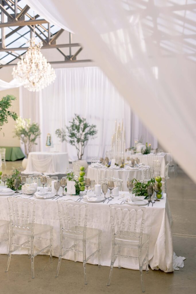A wide view of an indoor wedding reception at The Baumberhof featuring romantic white ceiling draping, a massive crystal chandelier, clear ghost chairs, and tables decorated with white linens and green fruit centerpieces.