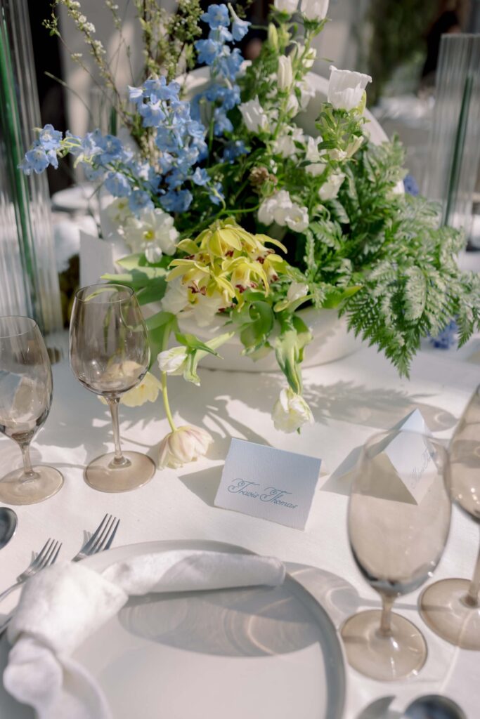 A close-up detail shot of a custom script wedding place card by Eden Hill Atelier styled on a sunlit reception tablescape with smoky tinted glassware and a blue and white floral centerpiece.⁠