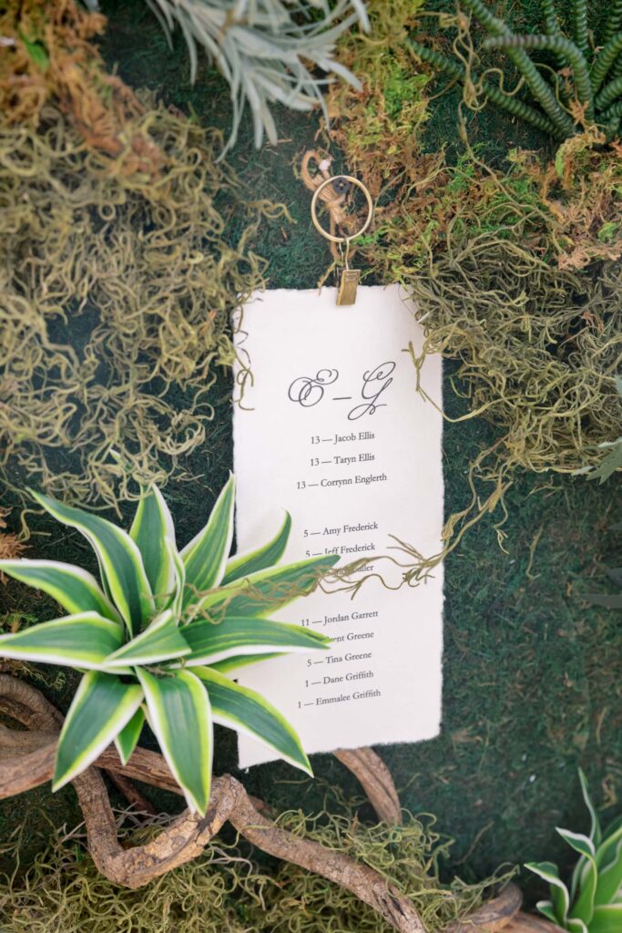 Close-up detail of a custom deckled-edge wedding seating chart card for letters E through G by Eden Hill Atelier, clipped to a green living moss wall beside a striped succulent.