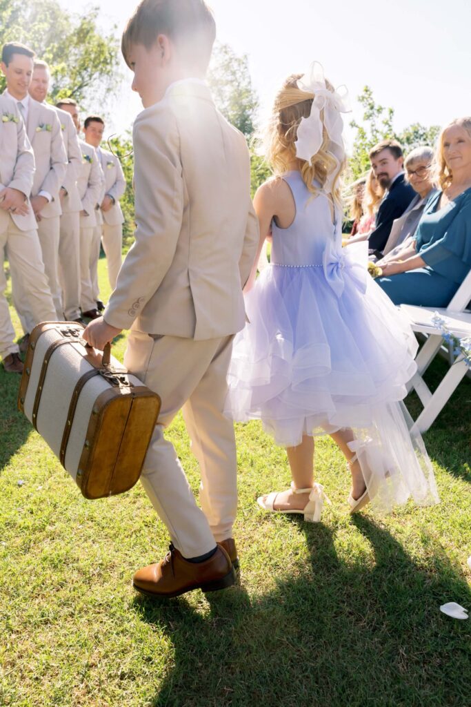 A ring bearer in a tan suit carrying a vintage suitcase walks down a sunlit grass aisle alongside a flower girl in a light purple tulle dress during an outdoor wedding ceremony.