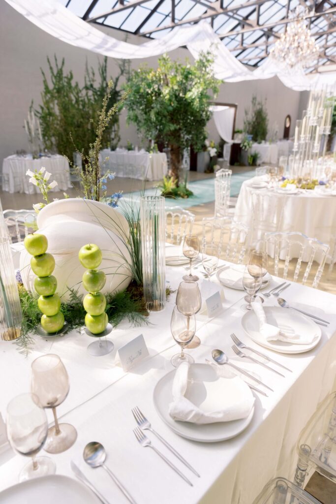 A bright indoor wedding reception tablescape featuring stacked green apples, a curved white ceramic vase centerpiece, smoky wine glasses, and custom tented place cards beneath a draped glass ceiling at The Baumberhof.