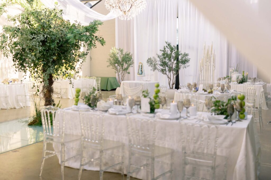 A wide horizontal photograph of an indoor wedding reception hall at The Baumberhof featuring tables with white linens, clear ghost chairs, unique green apple centerpieces, a large indoor tree, and white drapery backdrop.