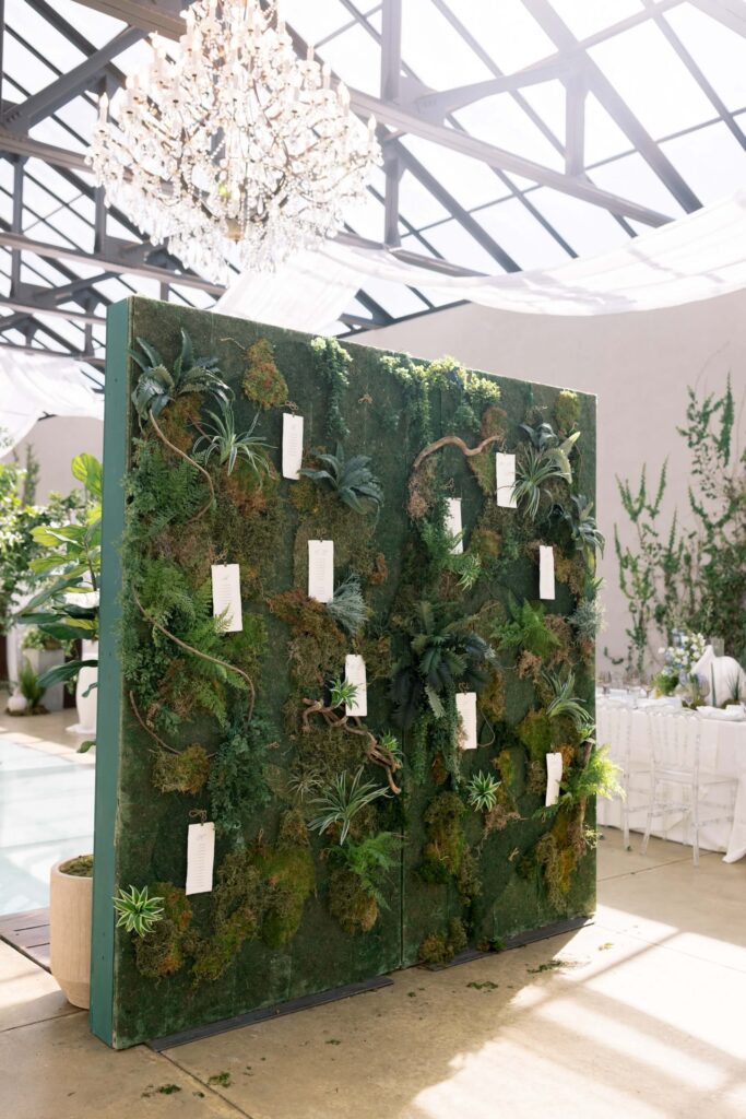 A large, free-standing green living moss wall seating display featuring suspended white escort cards by Eden Hill Atelier styled inside the glass conservatory at The Baumberhof.
