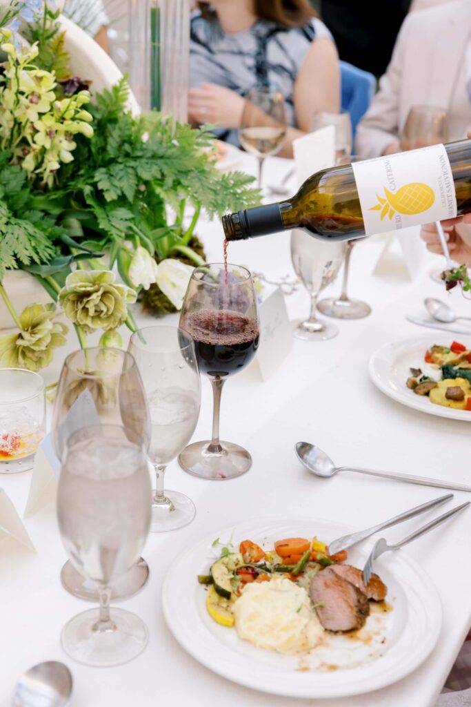 A close-up of a wedding reception table during a plated dinner service, showing red wine being poured into a tinted glass next to a custom script card by Eden Hill Atelier.