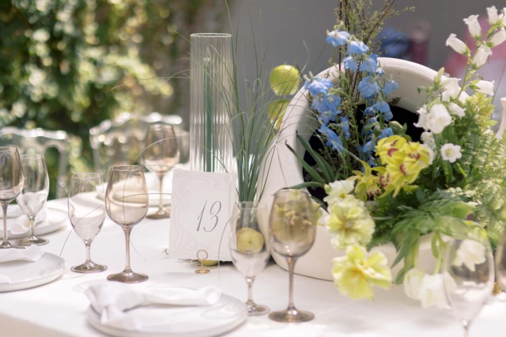 Horizontal shot of a fine art wedding reception tablescape featuring a custom Art Nouveau table number 13 card, a large white ceramic floral centerpiece, and elegant tinted wine glasses.⁠